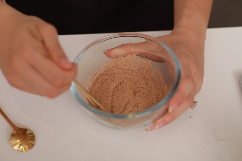 Hands mixing aromatic incense powders in a glass bowl with a wooden spoon